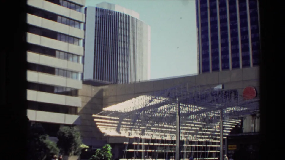 1981 New Zealand city plaza view with office towers, a geometric metal installation, and an unreadable red sign.