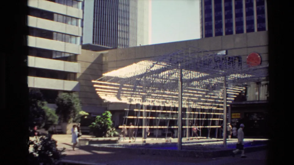 1981 New Zealand urban plaza scene with pedestrians passing a large geometric metal sculpture beneath high-rise buildings.
