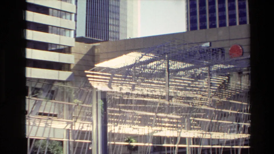 1981 New Zealand home-movie frame of a sunlit metal public-art structure in a modern urban plaza.