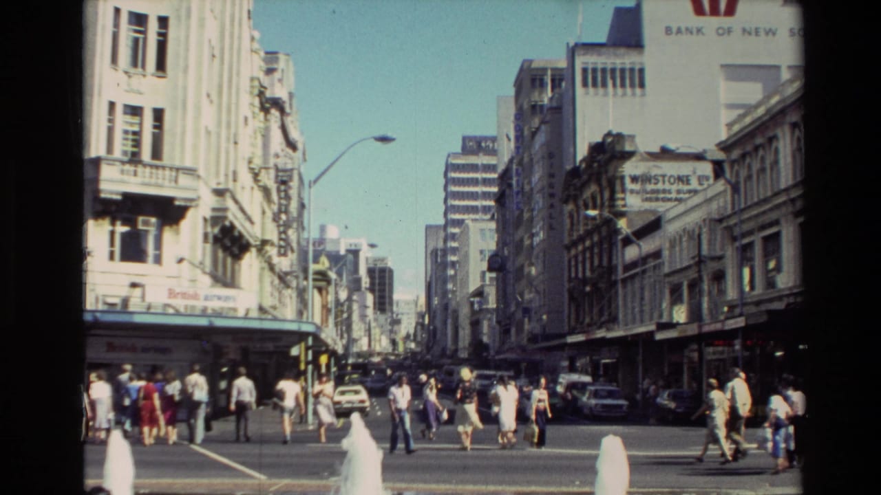 1981 New Zealand home-movie frame showing pedestrians crossing a bright urban street with cars, shopfronts and tall commercial buildings.