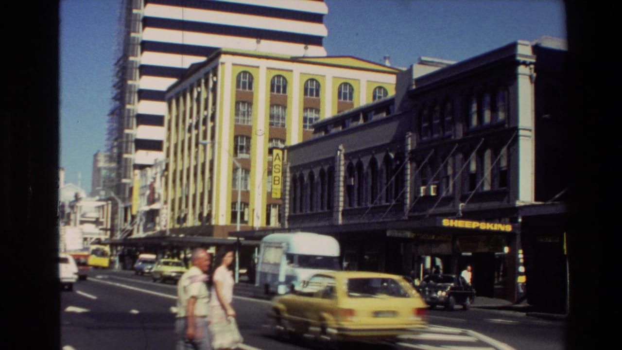 1981 New Zealand street view with a yellow car, pale blue van, pedestrians, Sheepskins signage and multi-story shop buildings.