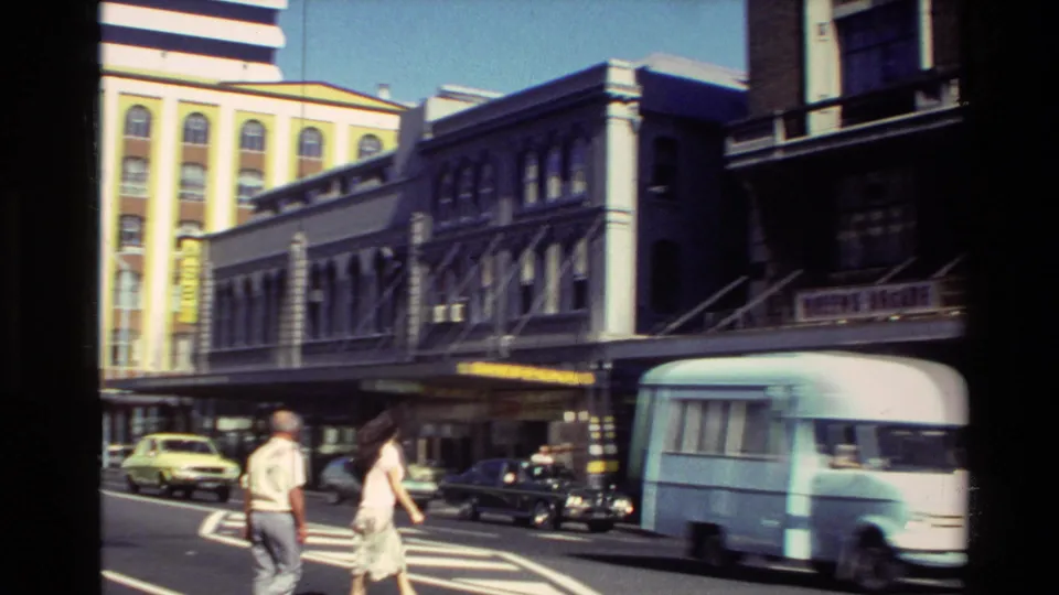 1981 New Zealand frame with pedestrians, a pale blue delivery van, classic cars and storefront signage along a city street.