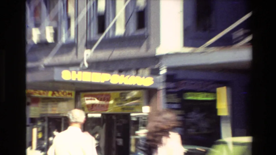 Soft 1981 New Zealand home-movie frame showing pedestrians and a bright Sheepskins storefront sign during a quick camera movement.