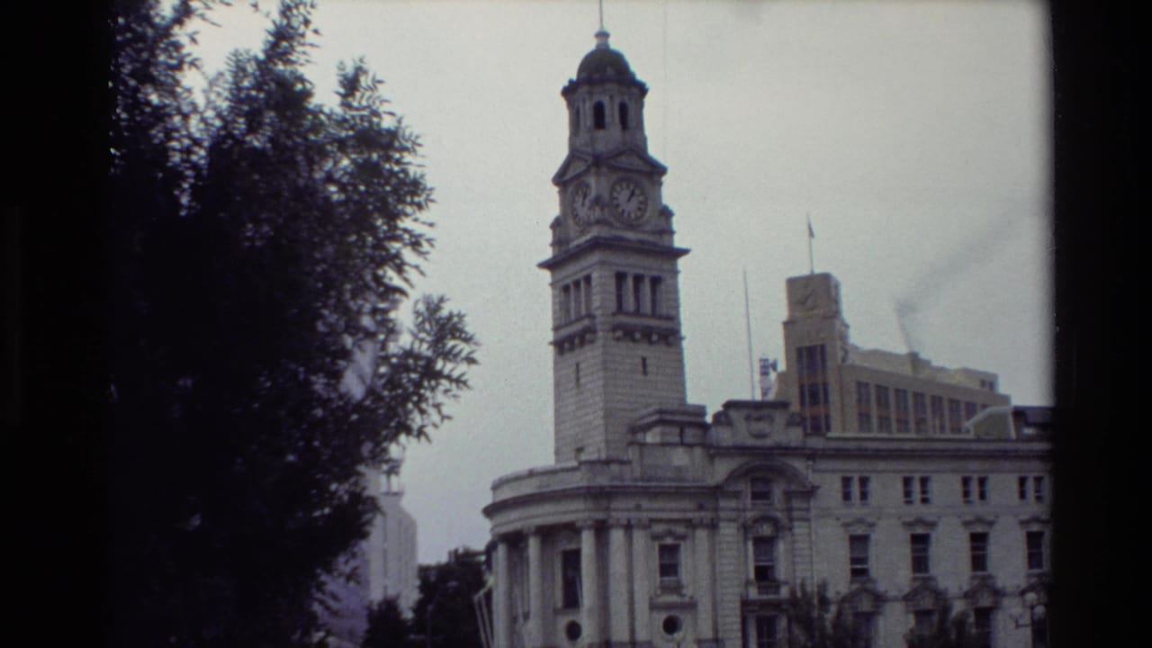 1981 New Zealand 8mm frame showing a historic clock-tower building viewed through trees in a public square.