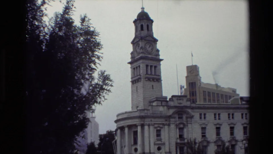 1981 New Zealand 8mm frame showing a historic clock-tower building viewed through trees in a public square.