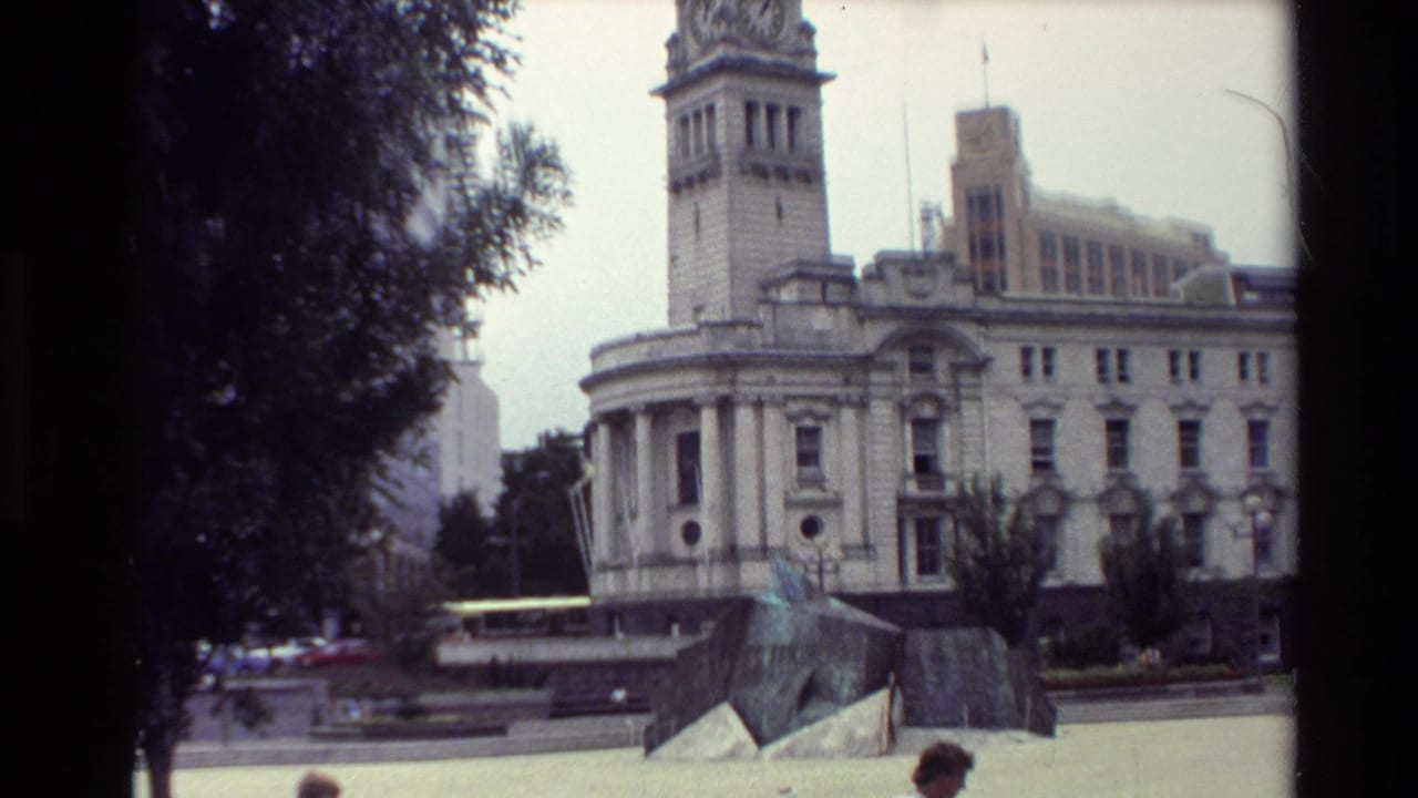 1981 New Zealand public square view with a clock-tower building behind a low sculptural monument and trees.