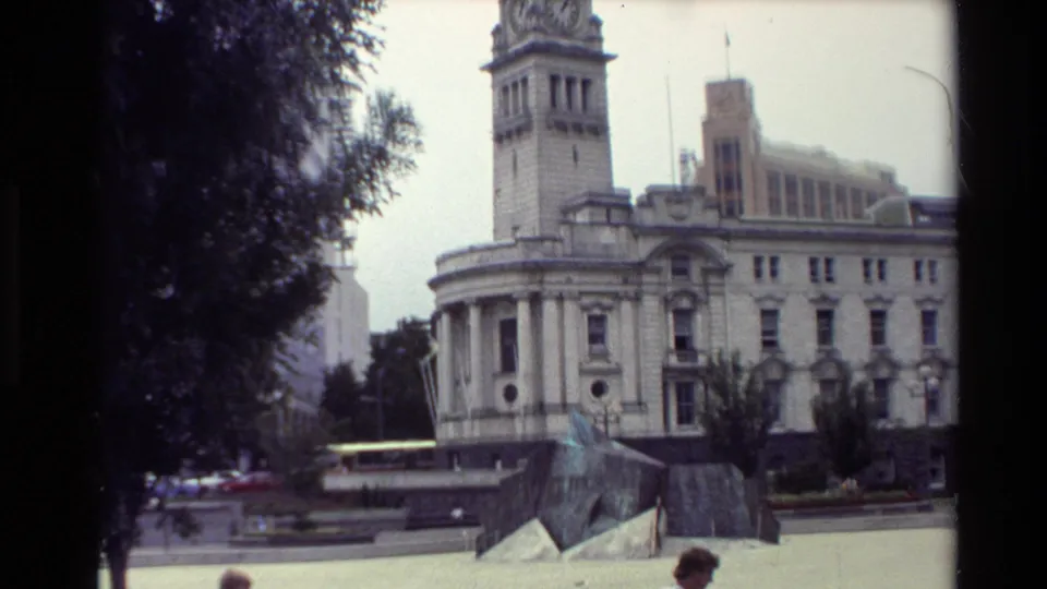 1981 New Zealand public square view with a clock-tower building behind a low sculptural monument and trees.