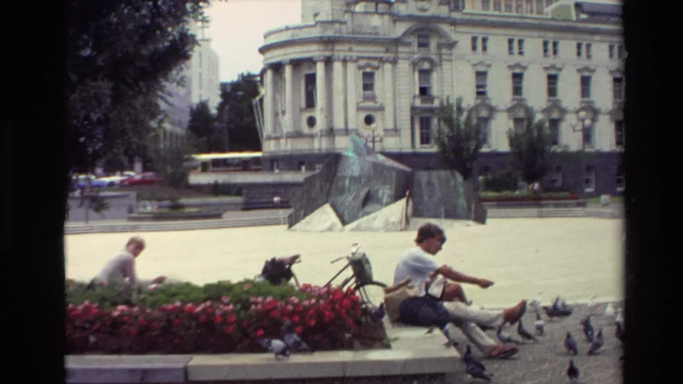 1981 New Zealand 8mm frame with seated visitors, bicycles, pigeons, flowers, and a historic clock-tower building beyond the plaza.