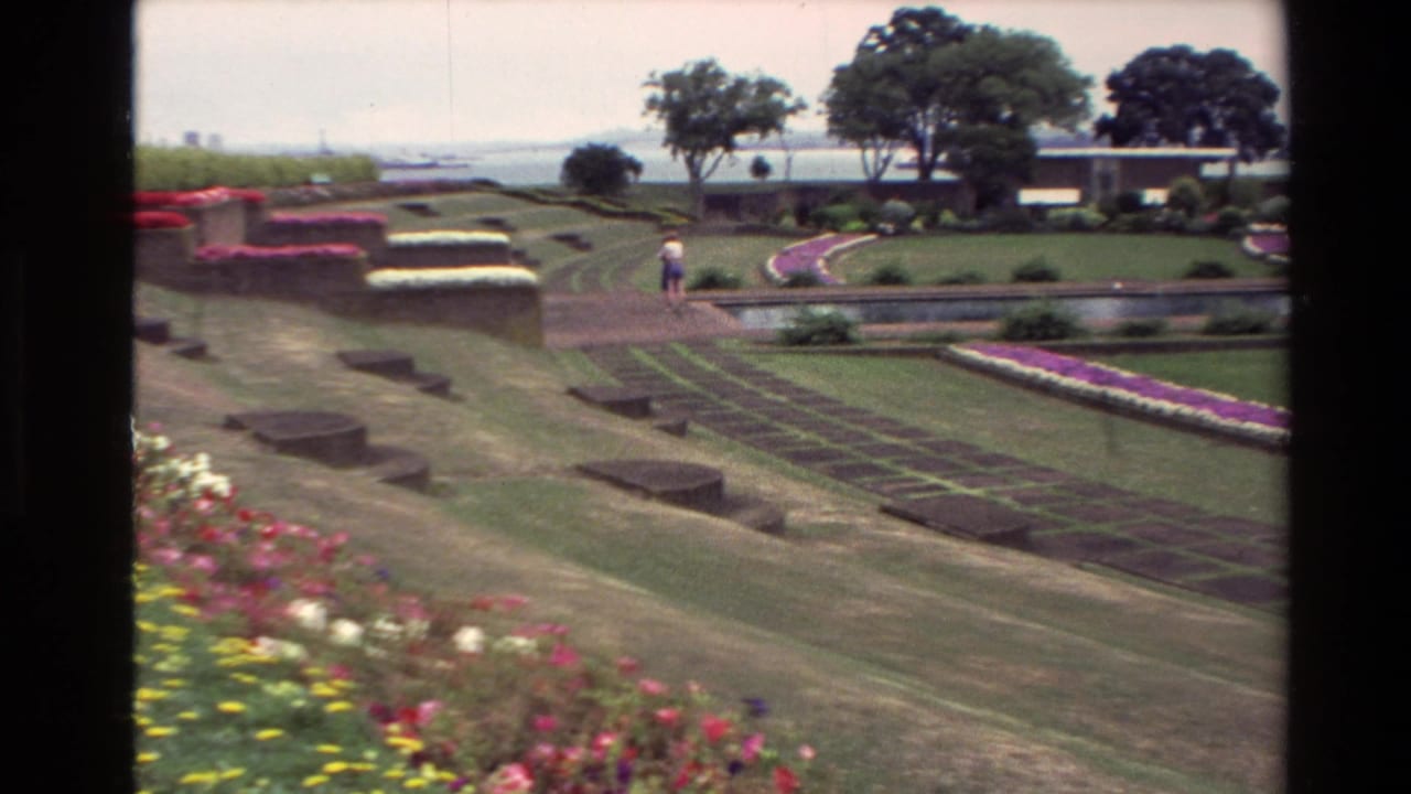 1981 New Zealand 8mm frame showing formal gardens, terraced lawns, colorful beds, a water feature, trees, and a distant walking visitor.