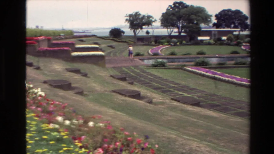 1981 New Zealand 8mm frame showing formal gardens, terraced lawns, colorful beds, a water feature, trees, and a distant walking visitor.