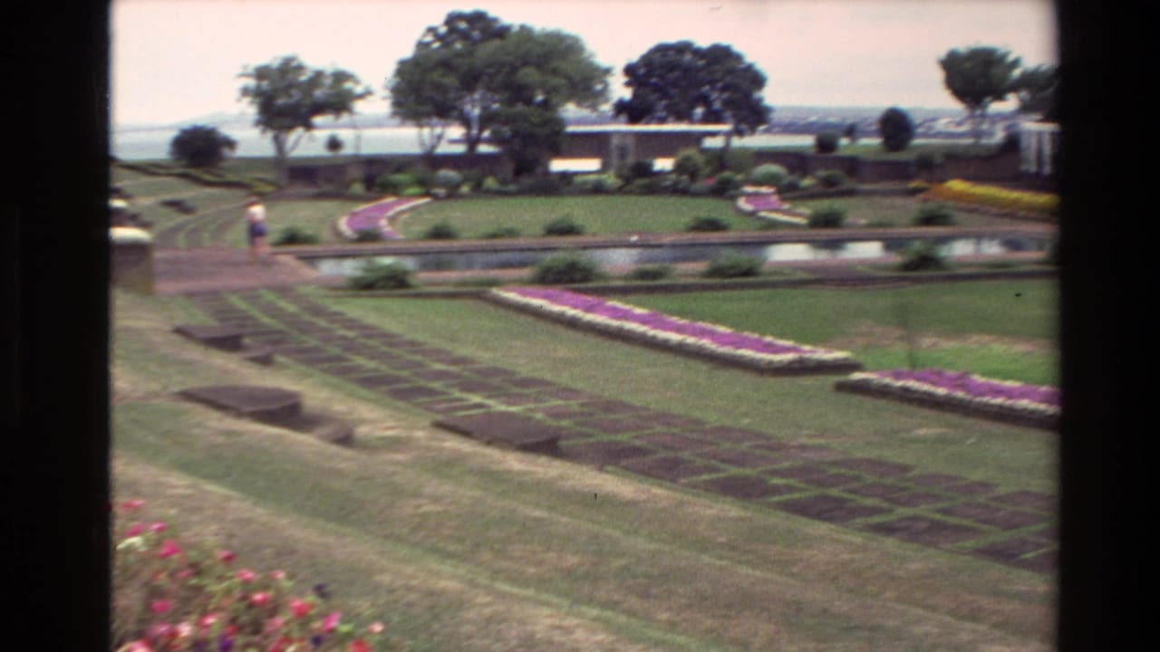 Formal garden slopes toward patterned beds and a water feature, with trees and distant water behind.