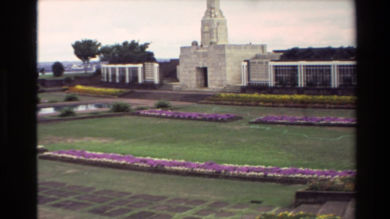 1981 New Zealand home-movie frame showing a stone memorial obelisk, white side structures, flowerbeds, lawns, and a reflective pool.