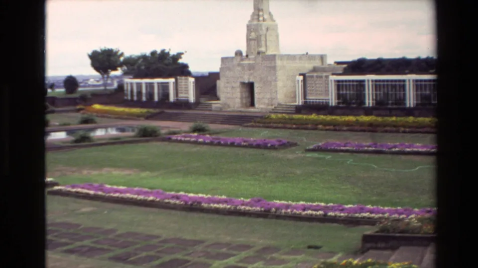 1981 New Zealand home-movie frame showing a stone memorial obelisk, white side structures, flowerbeds, lawns, and a reflective pool.