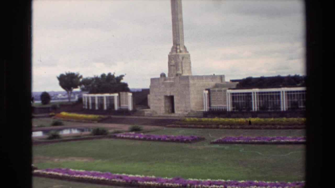 Visitor-shot 1981 New Zealand view of a tall memorial obelisk, formal garden beds, white structures, lawn, and cloudy sky.