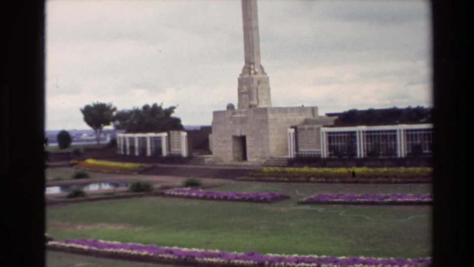 Visitor-shot 1981 New Zealand view of a tall memorial obelisk, formal garden beds, white structures, lawn, and cloudy sky.