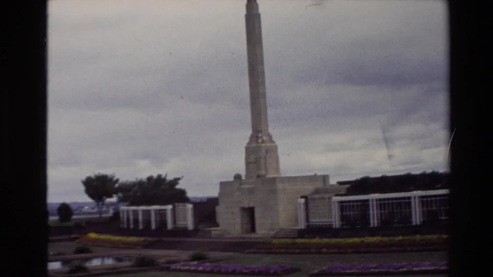 1981 New Zealand frame with a memorial obelisk rising above landscaped gardens, yellow and purple flowers, and overcast sky.