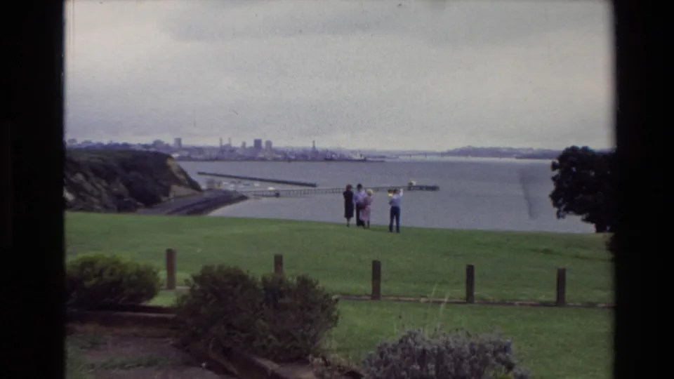 1981 New Zealand home-movie view of people on a grassy overlook facing a harbor, distant city buildings, shoreline, and cloudy sky.