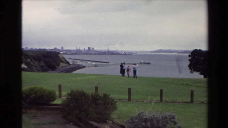1981 New Zealand home-movie frame of several adults at a coastal lookout, seen through dark frame edges with water and skyline beyond.