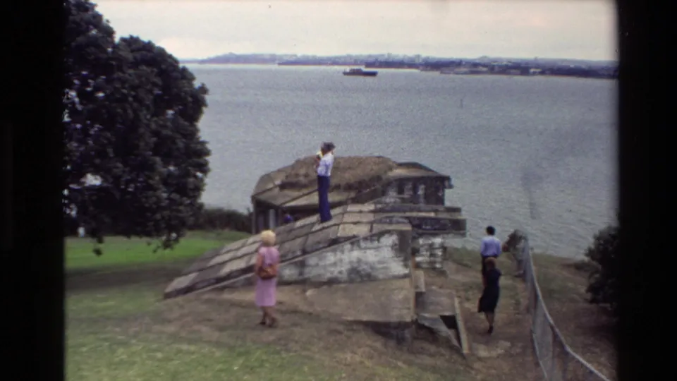 1981 New Zealand 8mm frame showing a woman in pink and other adults around a weathered stone structure overlooking a broad waterfront.