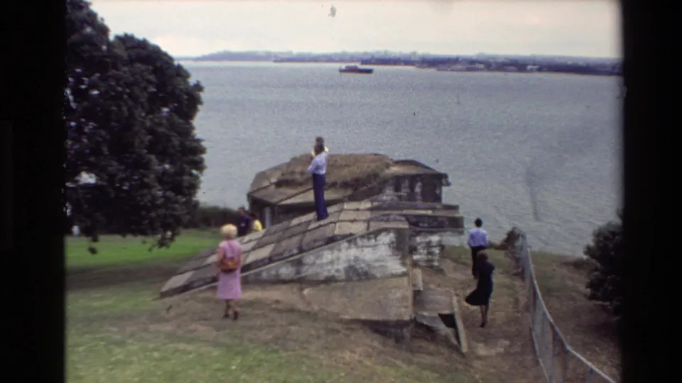 1981 New Zealand home-movie frame with adults near a weathered stone lookout structure, water behind them, and a ship in the distance.