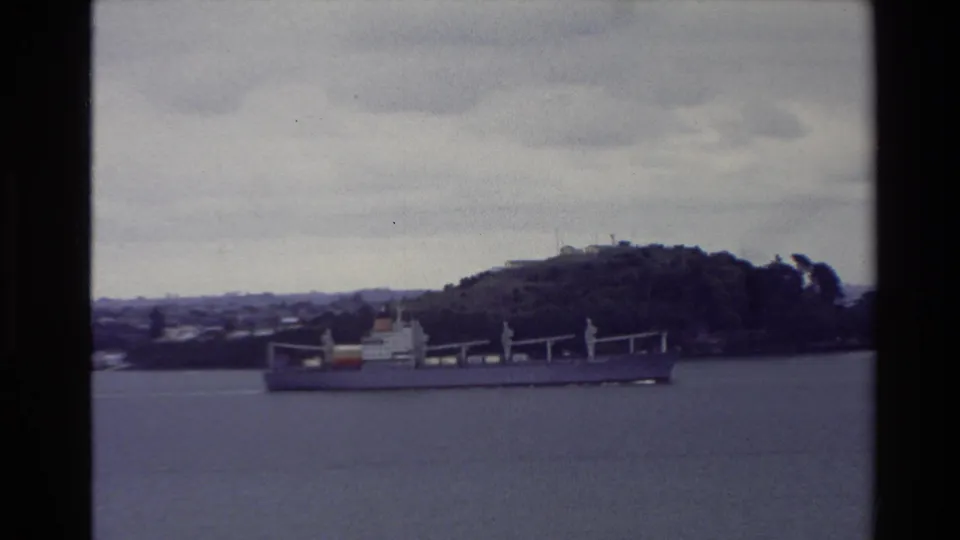 1981 New Zealand 8mm frame showing a cargo ship on harbor water with a wooded hill and overcast sky behind it.