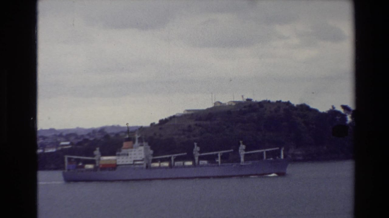 1981 New Zealand harbor view with a large cargo ship passing in front of a tree-covered hill, filmed on faded 8mm stock.