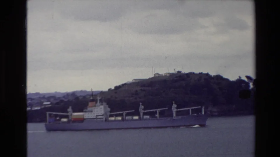 1981 New Zealand harbor view with a large cargo ship passing in front of a tree-covered hill, filmed on faded 8mm stock.