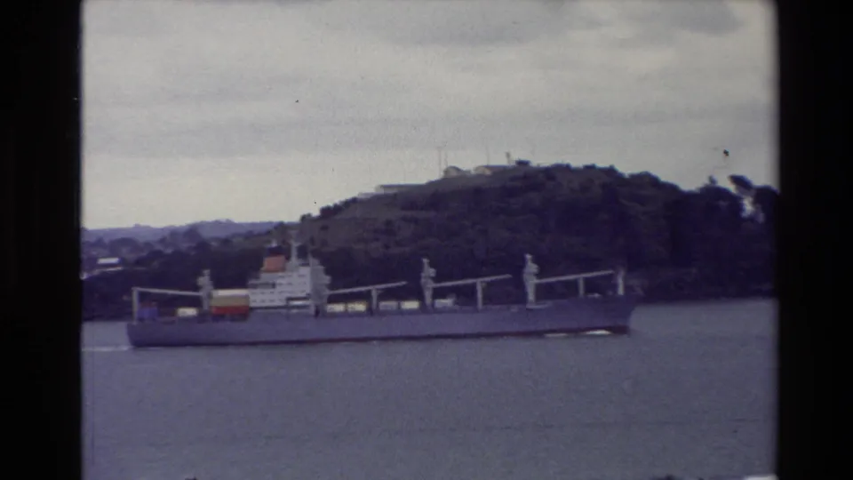 Faded 8mm New Zealand travel footage shows a cargo ship moving through harbor water near a wooded hillside.