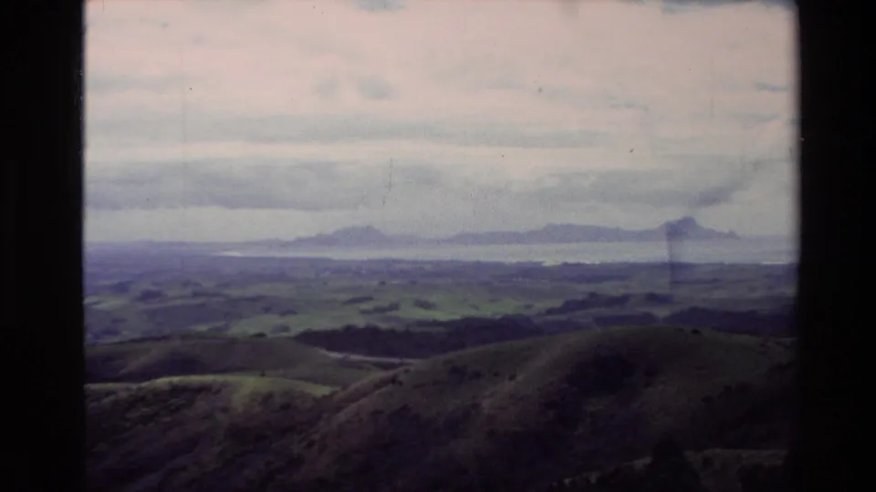 1981 New Zealand landscape view with rolling pasture, distant water or haze, and mountains under layered clouds.