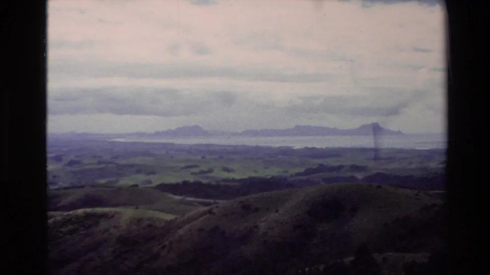 1981 New Zealand 8mm frame showing rolling green hills and distant mountains beneath a cloudy sky.