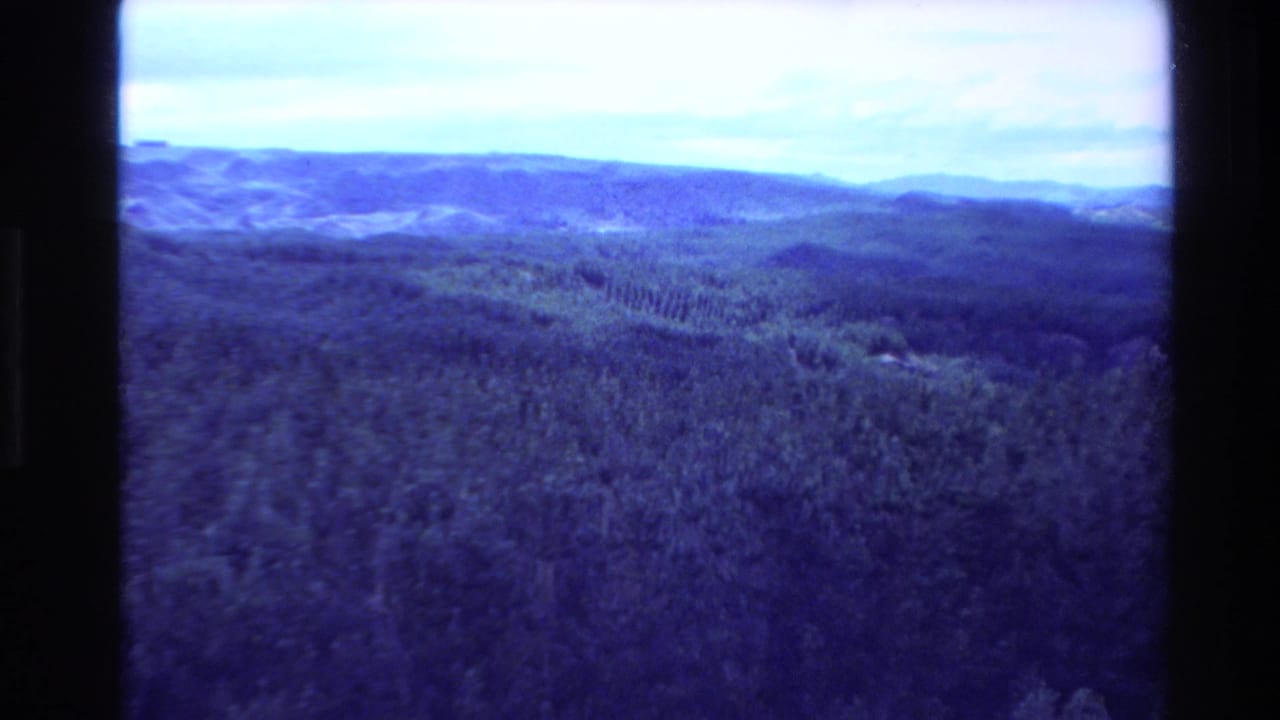 Blue-tinted aerial view of forested hills beneath a bright sky with dark 8mm frame edges.