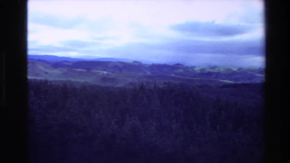 1981 New Zealand home-movie frame looking across rolling terrain, forested foreground, and cloud bands in a blue-tinted scan.