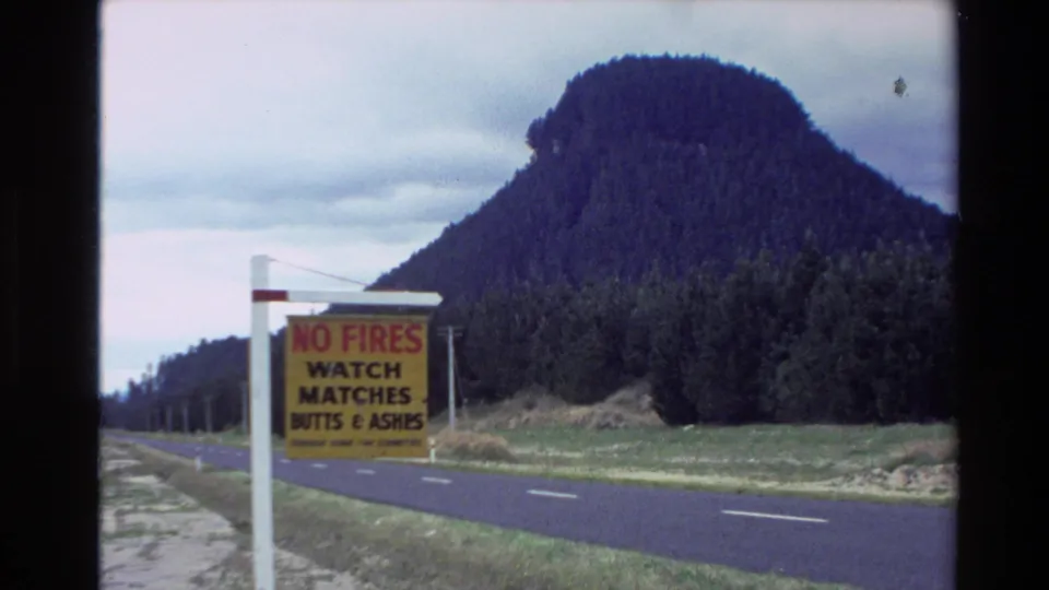 1981 New Zealand 8mm frame showing a roadside no-fires sign, empty road, grassy verge, and wooded mountain under cloud.