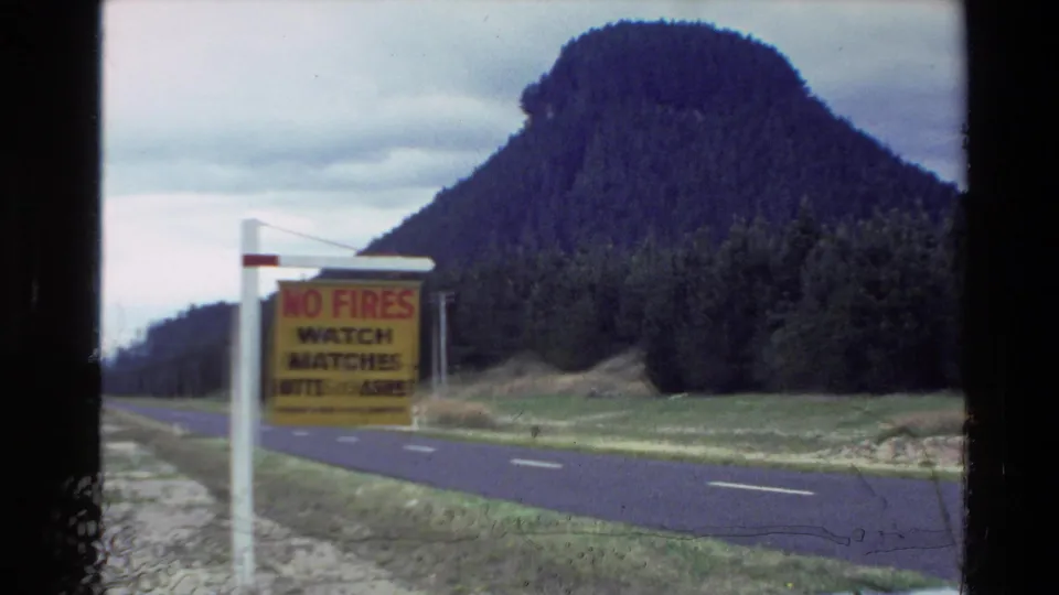 1981 New Zealand roadside view with a slightly blurred no-fires sign, two-lane road, grassy shoulder, and wooded mountain.