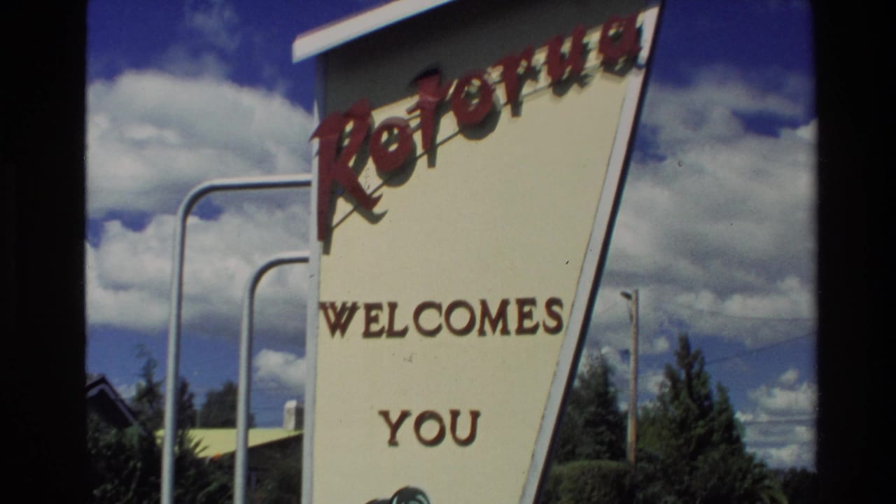 1981 New Zealand home-movie frame showing a Rotorua welcome sign with red lettering, greenery, and a cloud-filled sky.