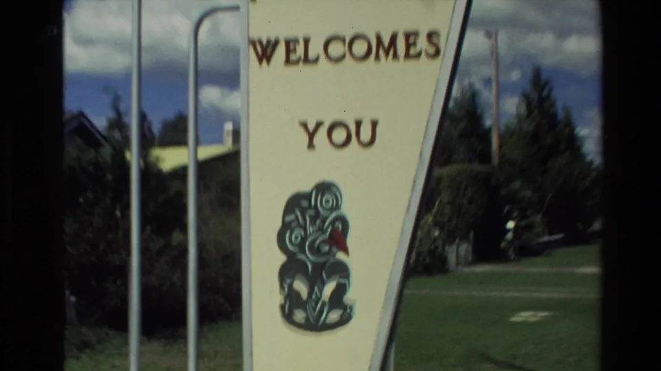 1981 Rotorua sign detail reading WELCOMES YOU, with a tiki-style graphic on a cream panel beside grass and trees.