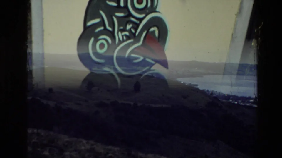 Home-movie transition frame combining the sign graphic with a distant New Zealand landscape of hills and water.