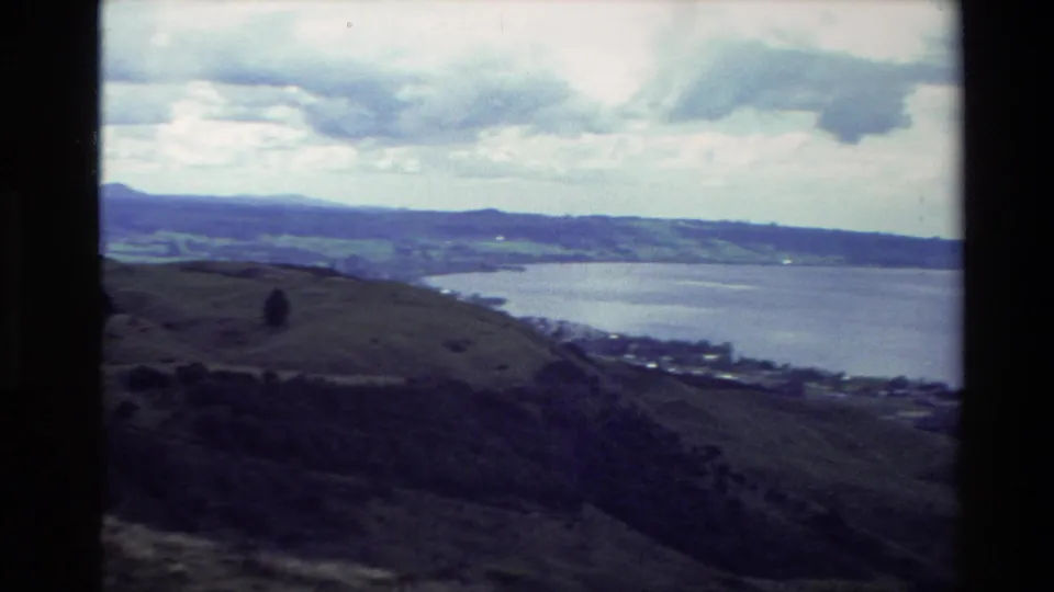 1981 New Zealand travel frame showing a broad lake view, rolling hills, distant shoreline, and heavy cloud cover.