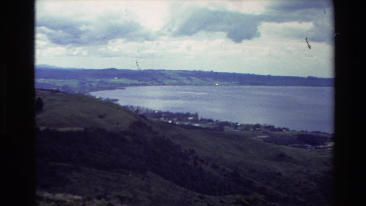 High view over a blue lake with rolling hills and shoreline farmland under clouds.