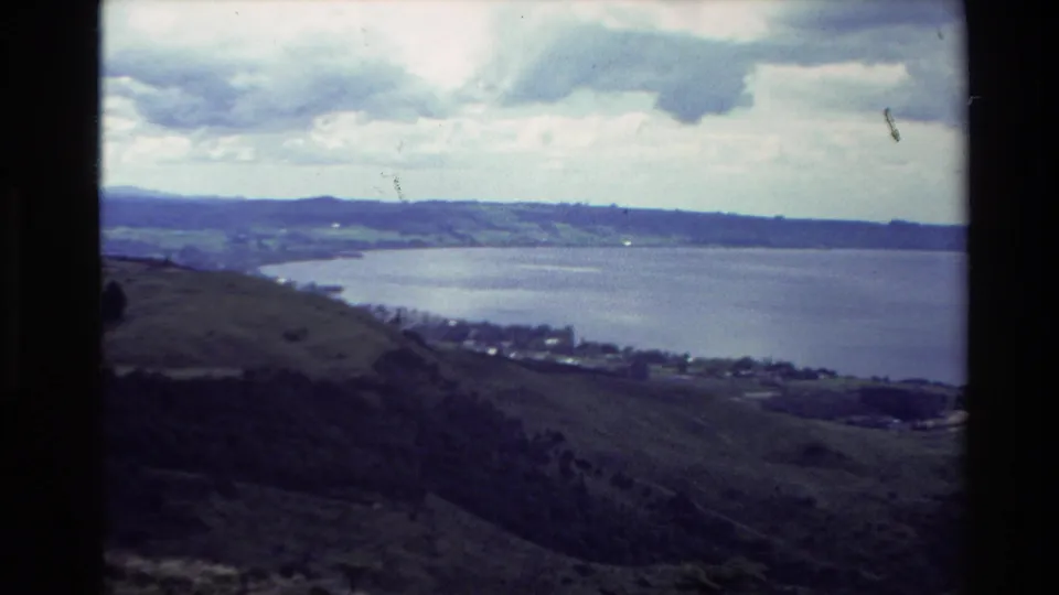 1981 New Zealand 8mm view from a hillside overlooking a blue lake, green slopes, and distant shoreline fields beneath a cloudy sky.