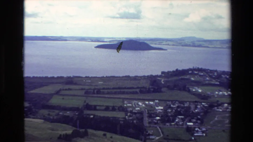 1981 New Zealand 8mm frame showing a small island in a broad lake, with divided green fields and village buildings along the shore.
