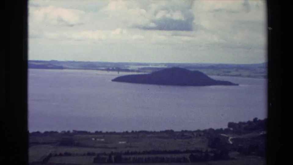 Vintage 8mm New Zealand lake view with a dark island across the water, foreground farmland, shoreline road traces, and clouded daylight.