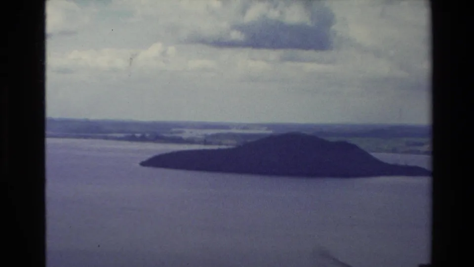 1981 New Zealand 8mm frame with the small lake island more prominent, open water around it, and hazy farmland and hills in the distance.