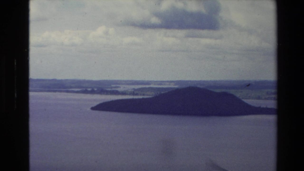 1981 New Zealand 8mm view of a dark, tree-covered island set in broad lake water beneath heavy clouds.