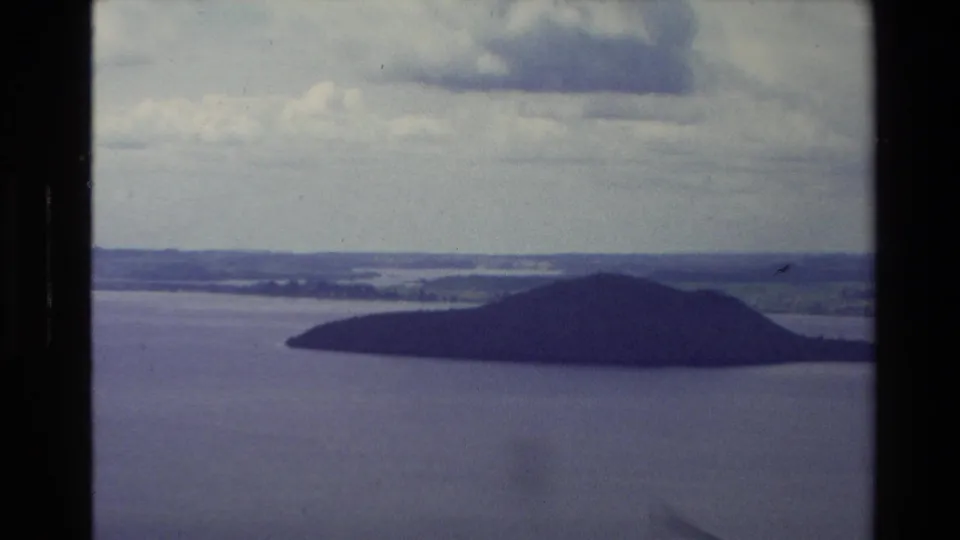 1981 New Zealand 8mm view of a dark, tree-covered island set in broad lake water beneath heavy clouds.