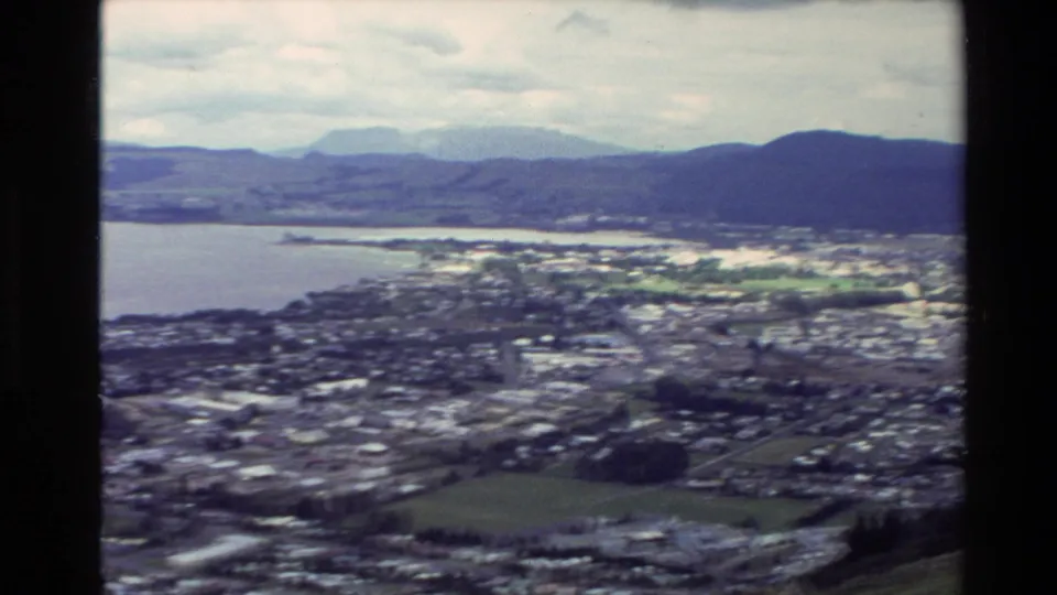Elevated 1981 New Zealand home-movie view across a lakeside city, shoreline, and distant hills under cloud cover.