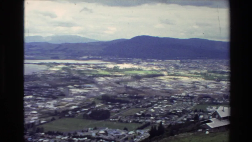 Soft 8mm frame showing a New Zealand lakeside city from above, with water, low buildings, and mountains beyond.