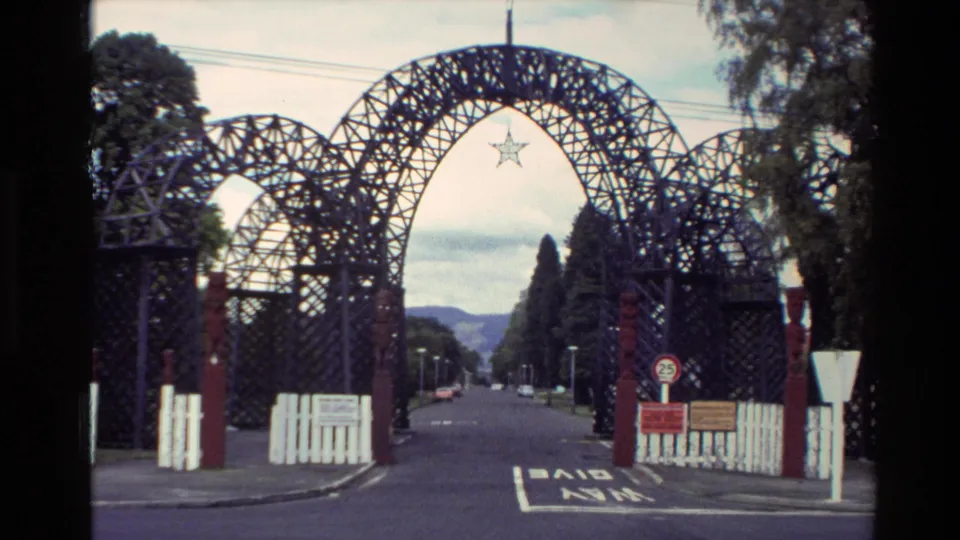 1981 New Zealand visitor-shot frame showing a decorative metal archway entrance over a road, with a star emblem, signs, trees, and distant water.