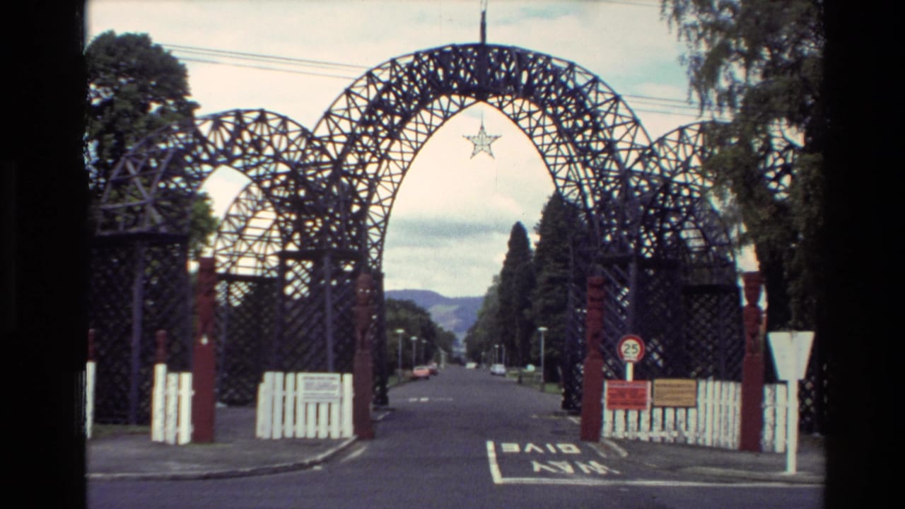 Latticework town entrance arch over a road with signs and white fencing.