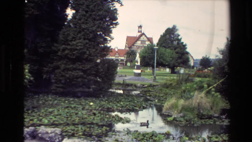 1981 New Zealand 8mm frame of a landscaped pond with lily pads and a duck, with trees and a Tudor-style building in the background.